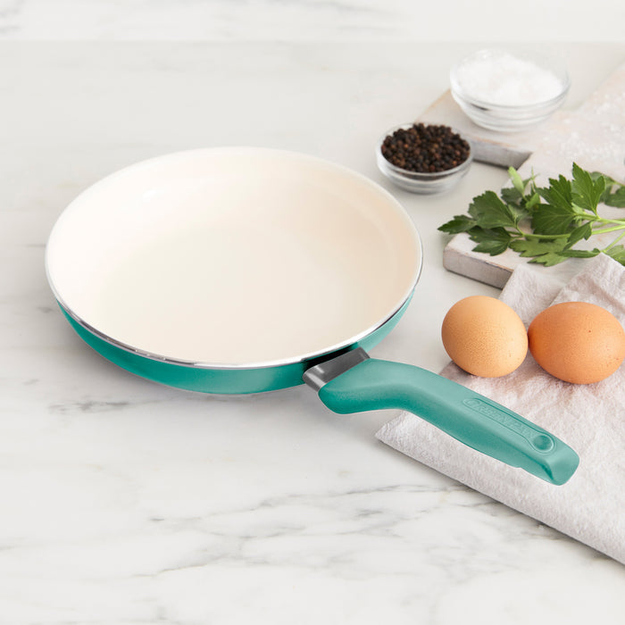 The Rio Ceramic Nonstick 8" Frypan with Spatula in turquoise rests on a marble countertop next to two eggs, fresh parsley, salt, peppercorns, a silicone spatula, and a folded white towel.