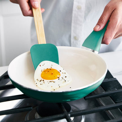 A person lifts a sunny-side-up egg topped with black pepper from the stove using the Rio Ceramic Nonstick 8" Frypan with Spatula in Turquoise.