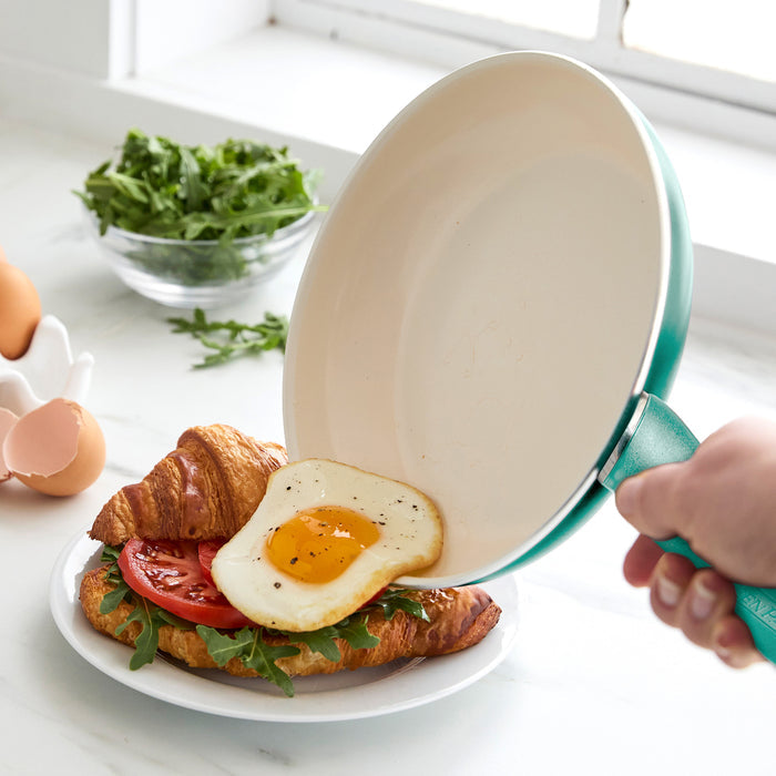 A person slides a fried egg from the Rio Ceramic Nonstick 8" Frypan with Spatula in Turquoise onto a croissant sandwich with tomato and arugula on a white plate. Nearby are a bowl of arugula, whole eggs, and eggshells on the counter.