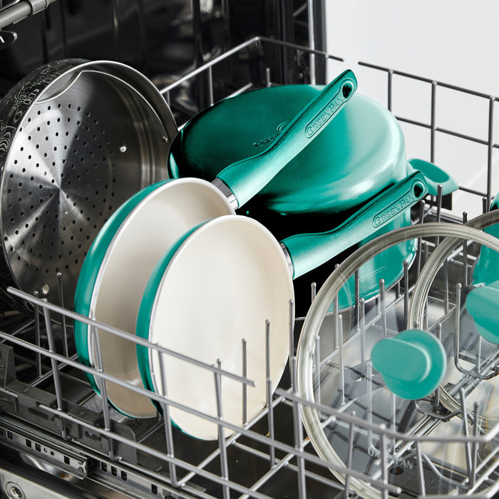 A dishwasher with a turquoise-handled Rio Ceramic Nonstick 8" Frypan with Spatula, white ceramic bowls, a metal steamer basket, glass lids, and more cookware neatly arranged in the top rack.