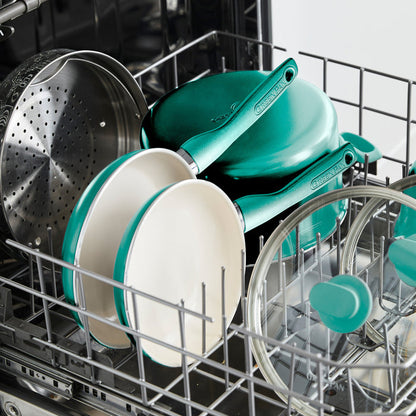 A dishwasher with a turquoise-handled Rio Ceramic Nonstick 8" Frypan with Spatula, white ceramic bowls, a metal steamer basket, glass lids, and more cookware neatly arranged in the top rack.