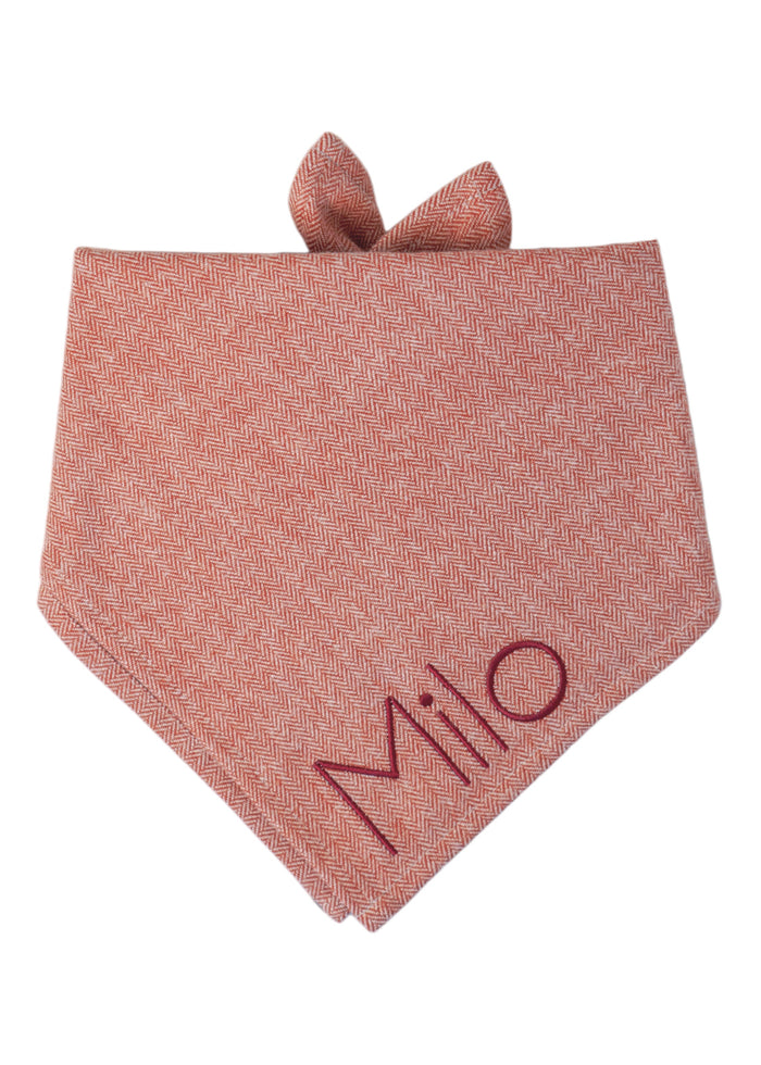 Classic herringbone dog bandana in muted red tones, tied at the top and embroidered with the name “Milo” stitched in deep red thread near the lower point.