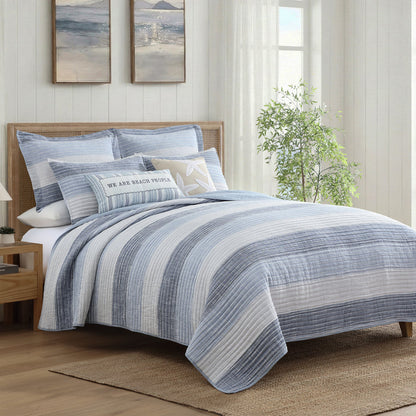 Sand Stripes Blue Quilt displayed on a bed with coastal decor and soft blue and white horizontal stripes.