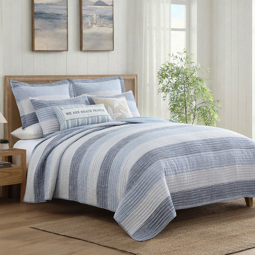 Sand Stripes Blue Quilt displayed on a bed with coastal decor and soft blue and white horizontal stripes.