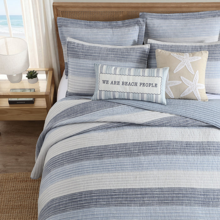 Sand Stripes Blue Quilt on a bed with beach-themed pillows and decor, showcasing a coastal-inspired look.