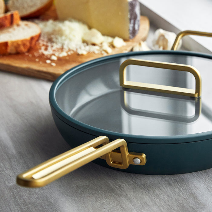 Venetian teal frypan with gold handles on a counter, bread and cheese blurred in the background.