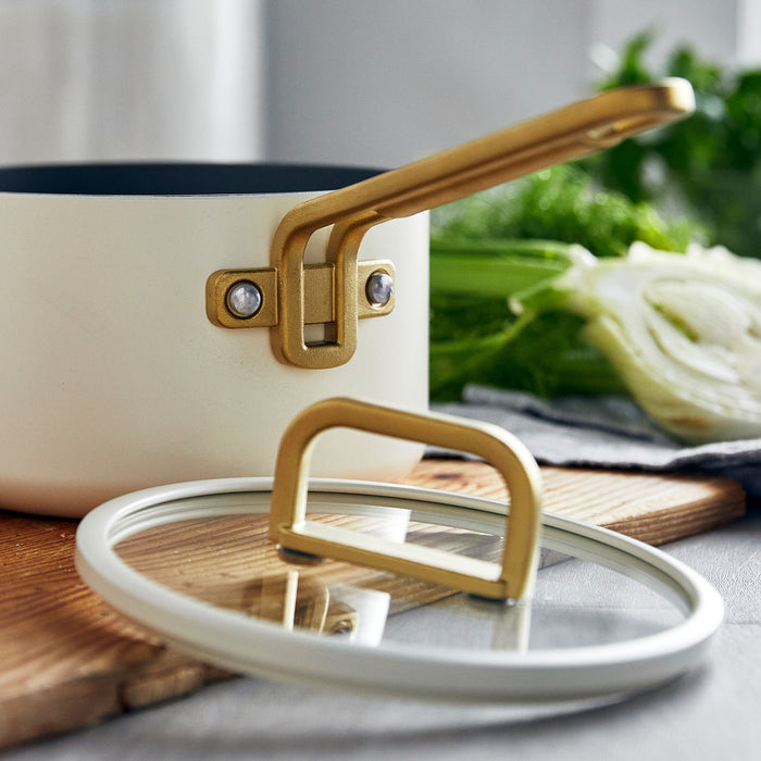 White ceramic nonstick saucepan with gold handle and lid, placed on wood next to assorted vegetables. 2-quart size.