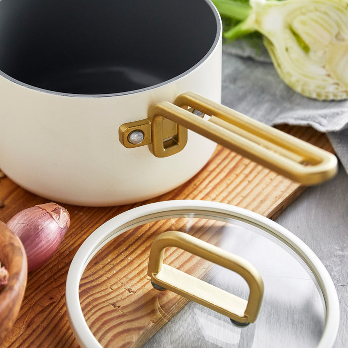 White ceramic nonstick saucepan with gold handle and lid next to shallot and fennel. 2-quart size.