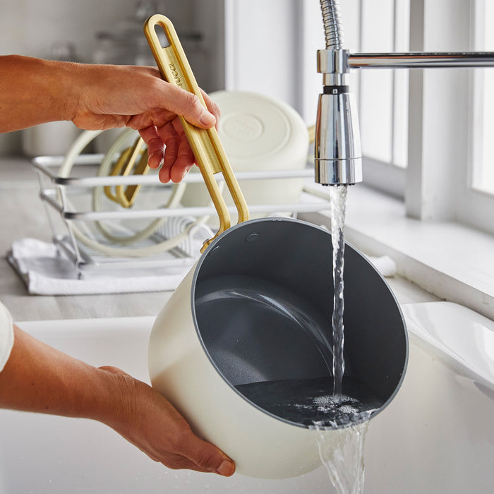 White ceramic 2-quart saucepan with lid being washed under running water in a kitchen sink.