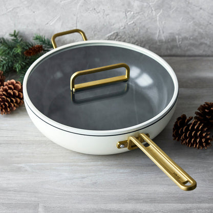White ceramic nonstick pan with gold handles and glass lid on a wooden surface, pinecones nearby.