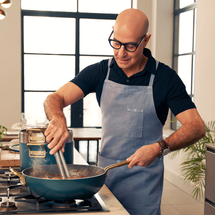 Man in glasses and apron stirring food in a white ceramic nonstick pan in a bright kitchen.