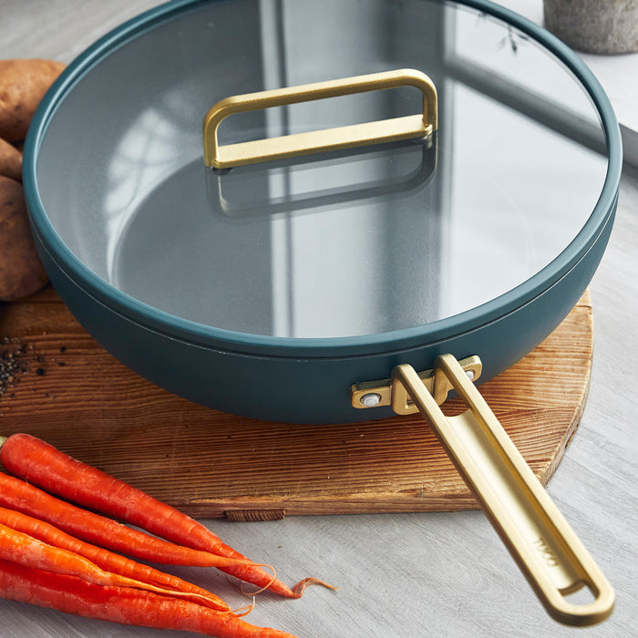 Venetian teal 6.5-quart pan with a glass lid and gold handle on a wooden board.