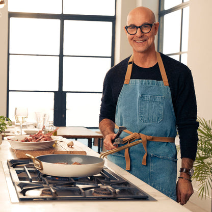 Man in glasses and denim apron using Stanley Tucci 11-piece cookware set in a modern kitchen.