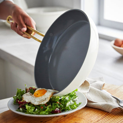 A hand slides a sunny-side-up egg from a white ceramic nonstick frying pan onto a salad plate.
