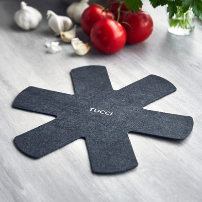 Black felt pan protector with TUCCI text next to tomatoes, garlic, fresh herbs, and a ceramic nonstick frypan set.