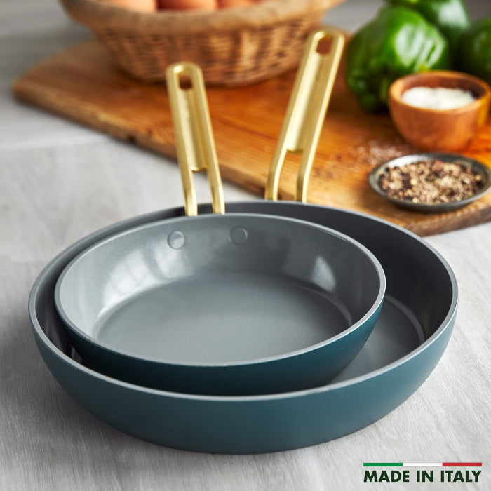 Two Stanley Tucci™ Ceramic Nonstick 8" and 10" Frypans in Venetian Teal with gold handles are stacked on a kitchen counter. Nearby are eggs, a green pepper, seasoning, and a bowl. Text reads MADE IN ITALY. PFAS-free cookware.