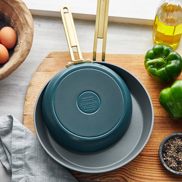 Two Stanley Tucci™ Ceramic Nonstick 8" and 10" Frypans in Venetian Teal sit on the counter near a green bell pepper, small bowl of pepper, olive oil, eggs in a wooden bowl, and gray cloth—perfect for PFAS-free cooking.