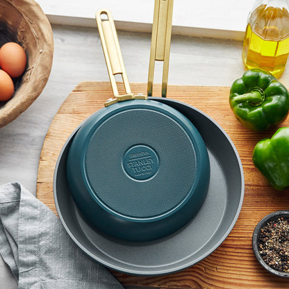 Two Stanley Tucci™ Ceramic Nonstick 8" and 10" Frypans in Venetian Teal sit on the counter near a green bell pepper, small bowl of pepper, olive oil, eggs in a wooden bowl, and gray cloth—perfect for PFAS-free cooking.