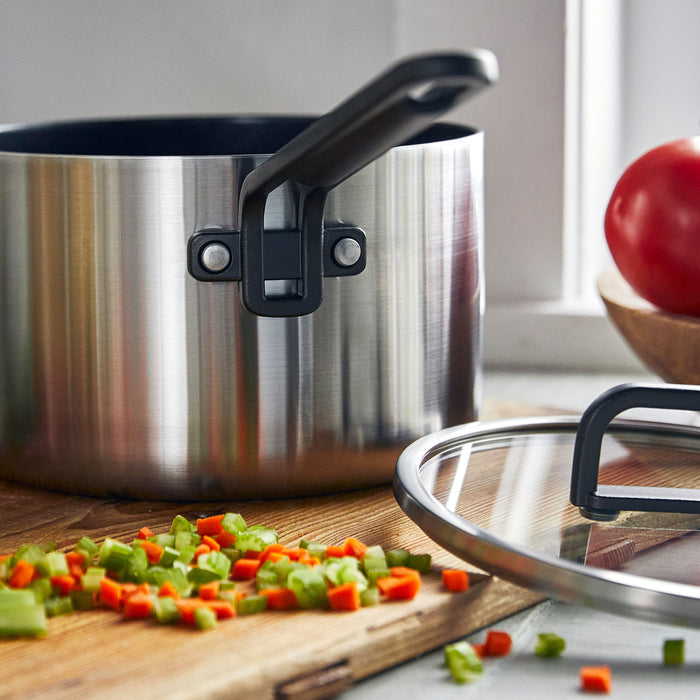 Black nonstick saucepan with lid on a wooden board, next to chopped carrots, celery, and a tomato.