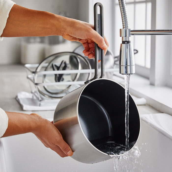 Person rinsing a stainless steel saucepan with black ceramic interior under a kitchen faucet.