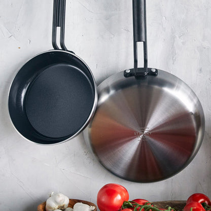 Two stainless steel nonstick frypans, one facing up and one down, with tomatoes and garlic on a neutral background.