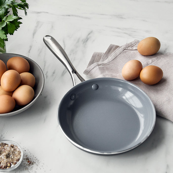 Ceramic nonstick 8-inch frypan next to brown eggs, fresh herbs, gray napkin, and a small dish of seasoning.
