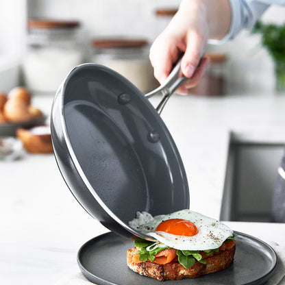 A hand slides a fried egg from a gray ceramic nonstick frypan onto toast with greens on a gray plate.