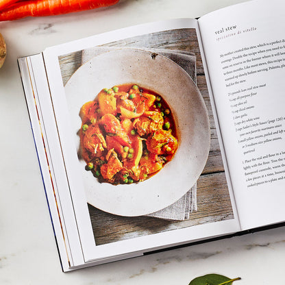 The Tucci Cookbook open to Italian recipes, next to a white bowl of veal stew and vegetables on a table.