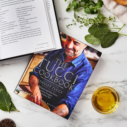 Tucci ceramic frypan set with two pans displayed on a table beside herbs, olive oil, and an open cookbook.