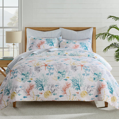 Bed with floral-patterned bedding in a bright room with plants and a lamp.